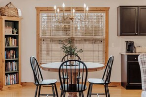 A dining area with a round table, four chairs, a chandelier, a window with blinds, a bookshelf, and a kitchen area with cabinets and a coffee maker. - Cozy Pines Cabin | Gated Community | Pool + Golf Course | Near SDC (Branson West)