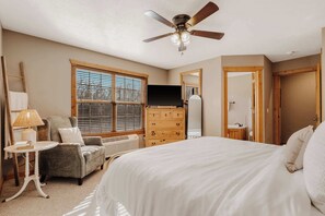 Quarto com cama, penteadeira, cadeira, luminária, televisão e janela com persianas.