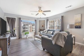 A modern living room with a grey sofa, a ceiling fan, and large windows. - Brookside Duo | Peaceful Views! (Branson)