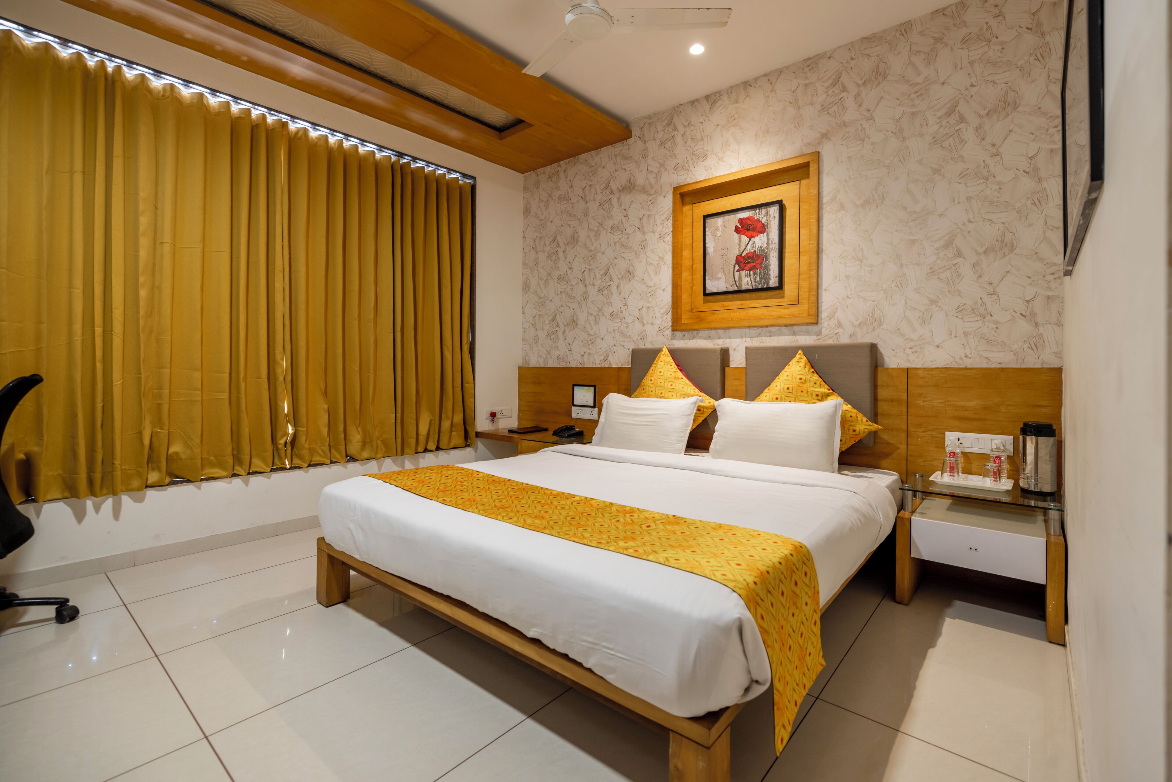 Une chambre d’hôtel avec un grand lit, une table de chevet, une chaise, un tableau encadré au mur et une fenêtre avec des rideaux.