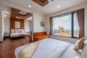 A hotel room with a large bed, a sofa, a ceiling fan, a window with a view of a cityscape, and a wall-mounted air conditioner. - Manek the Ocean View (Dwarka)