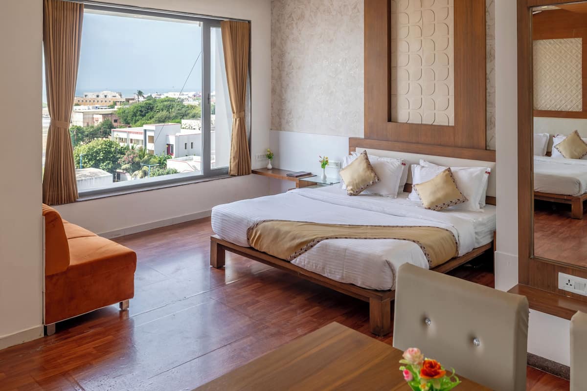 A hotel room with a bed, a dining table with a vase of flowers, a view of the city through a large window, and two hanging pendant lights.