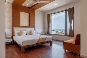 A bedroom with a bed, a nightstand, a chair, a large window, and a ceiling fan. - Manek the Ocean View (Dwarka)