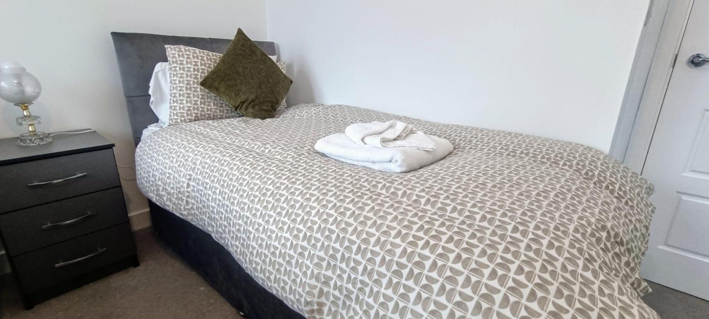 A neatly made bed with a patterned bedspread, a folded towel, and a single pillow.