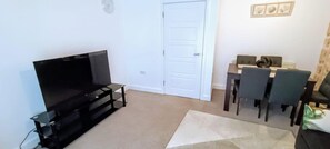 A living room with a flat-screen TV on a stand, a dining table with chairs, and a door. - Cozy Peace in Sittingbourne (Sittingbourne)