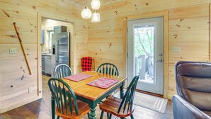 Interior de una cabaña de madera con una mesa de comedor, sillas, un refrigerador y una puerta que da al exterior.