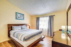 Una cama bien hecha con una colcha a rayas, un cabecero de madera, una mesita de noche con una lámpara, una ventana con cortinas y un cuadro enmarcado en la pared.