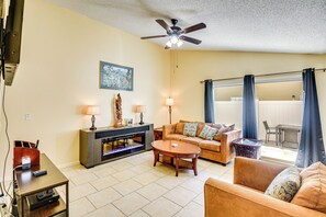 A living room with a sofa, coffee table, TV, fireplace, and a ceiling fan. - Palm Coast Home w/ Hot Tub - 3 Mi to Beaches! (Palm Coast)
