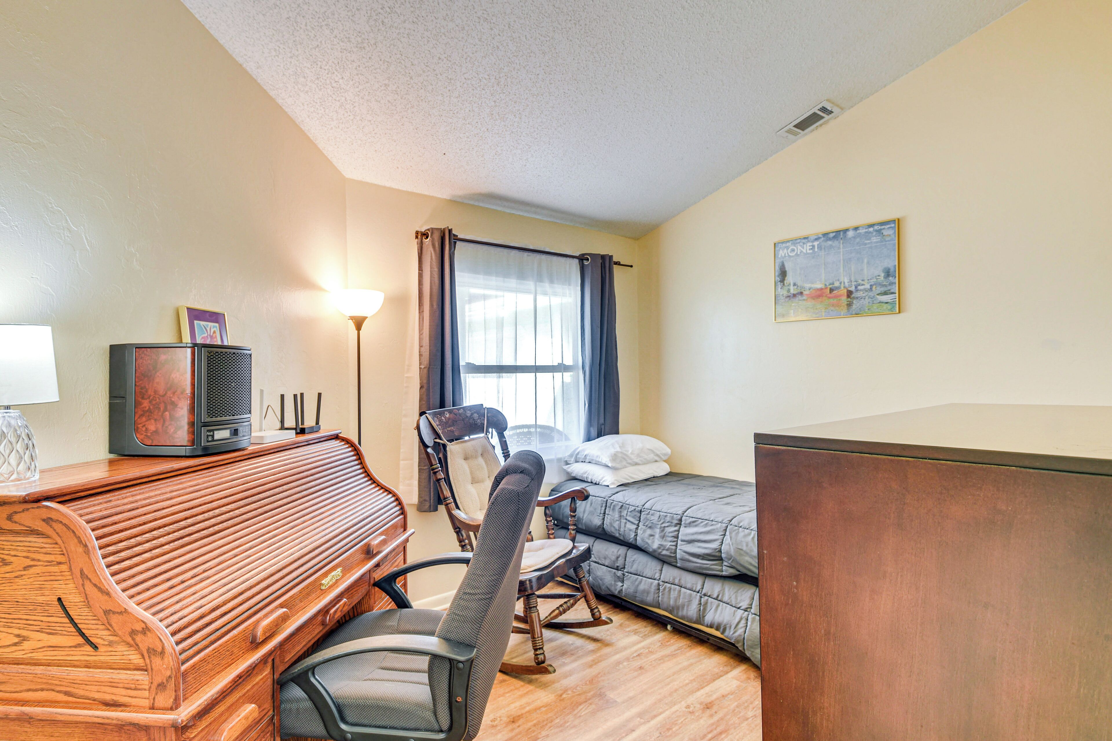 A compact hotel room with a wooden piano, a rocking chair, a bed with a blue blanket, and a painting on the wall.
