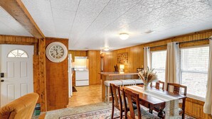 A cozy kitchen with wooden walls, a large clock, and a dining area with a table and chairs. - Heber Springs Cabin w/ Deck: 3 Mi to Sugarloaf Mtn (Heber Springs)
