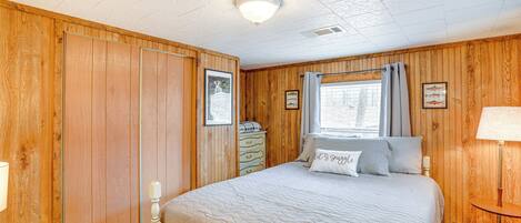 A bedroom with wooden paneling, a bed with a quilt, a dresser, a window with curtains, and a bedside lamp.