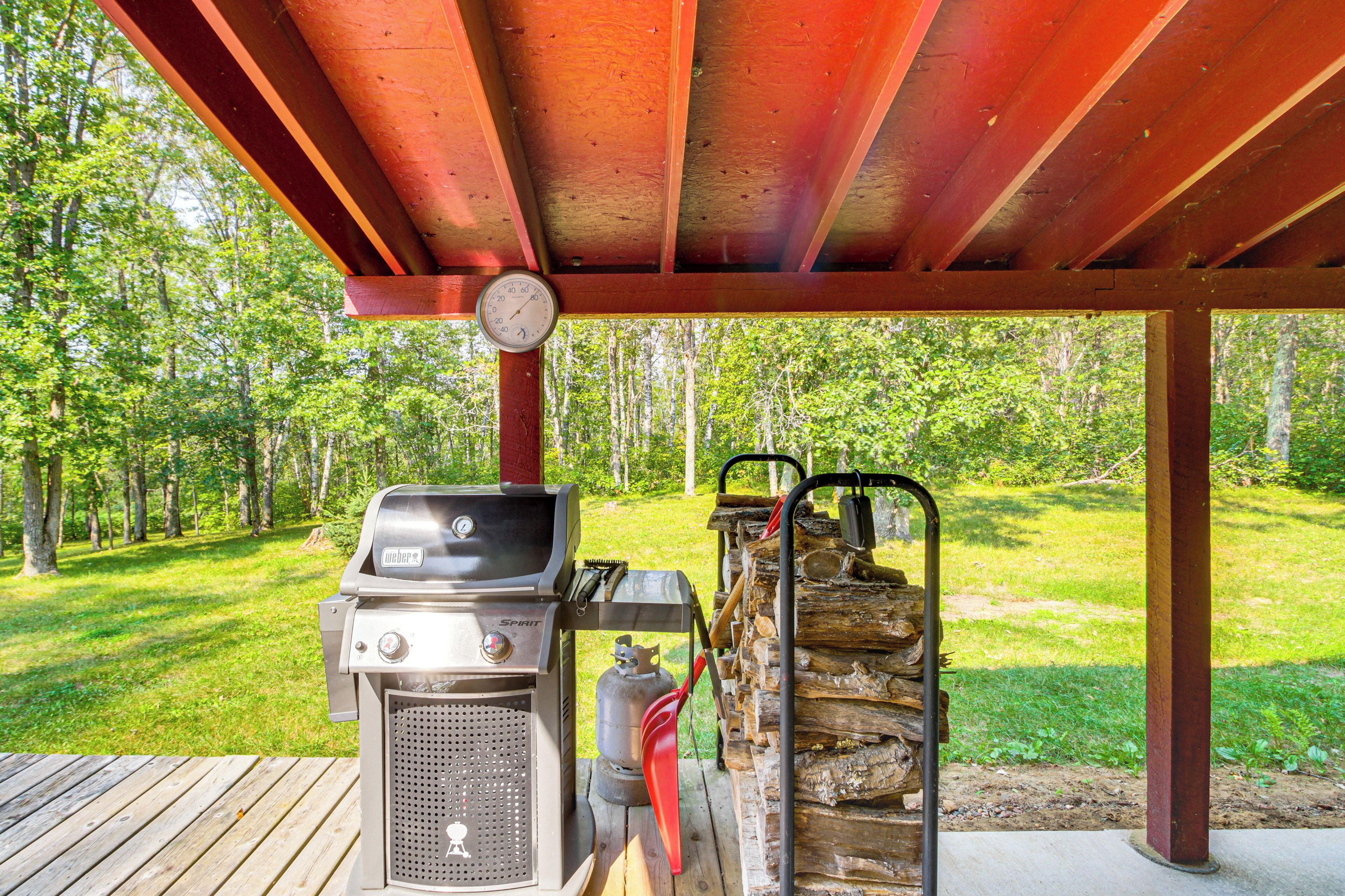 Ein überdachter Grillbereich mit einem Spirit-Grill, einer Propangasflasche und einem Holzstoß unter einem Holzdach.