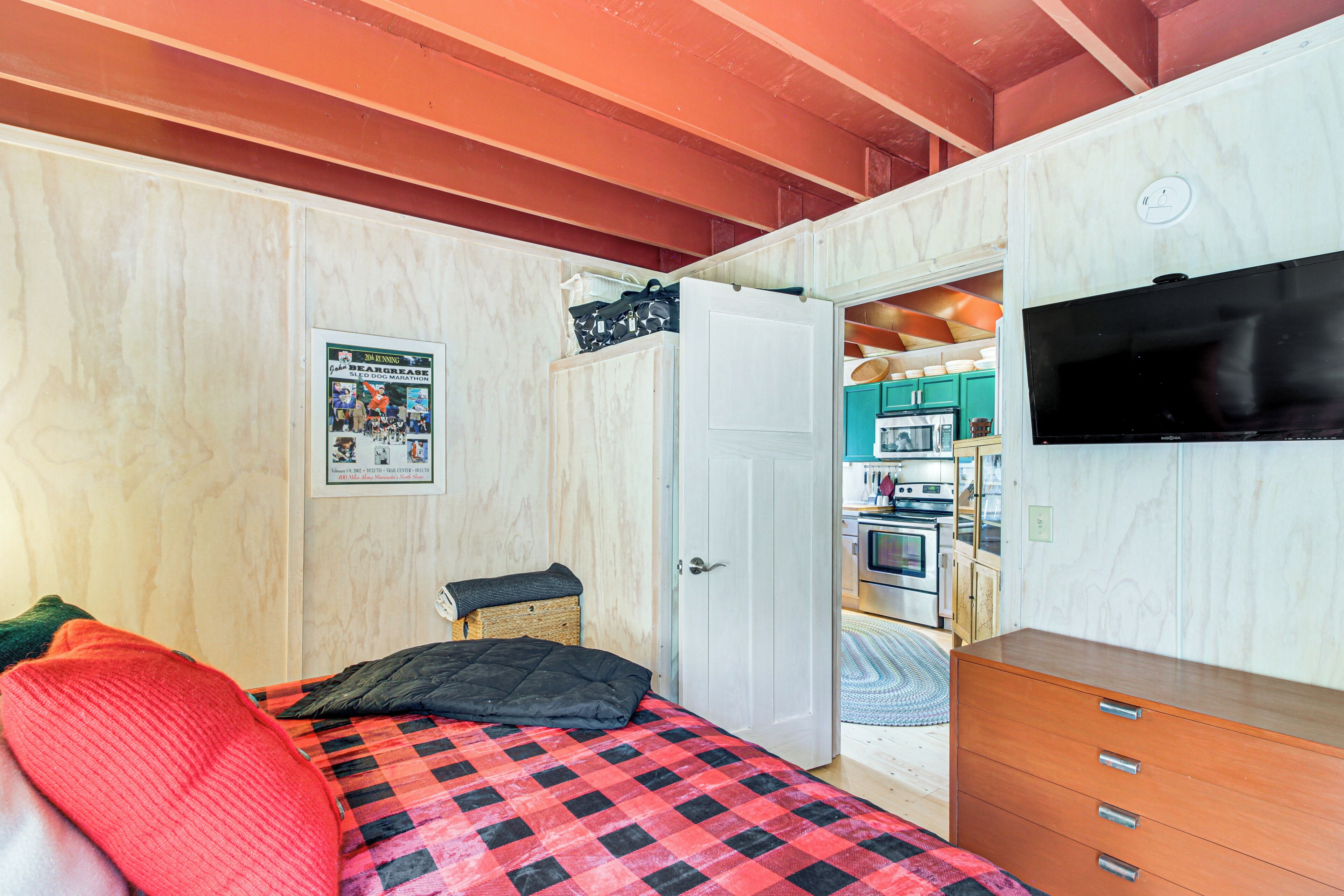 A bedroom with a bed, a dresser, a television, and a door leading to a kitchen area.