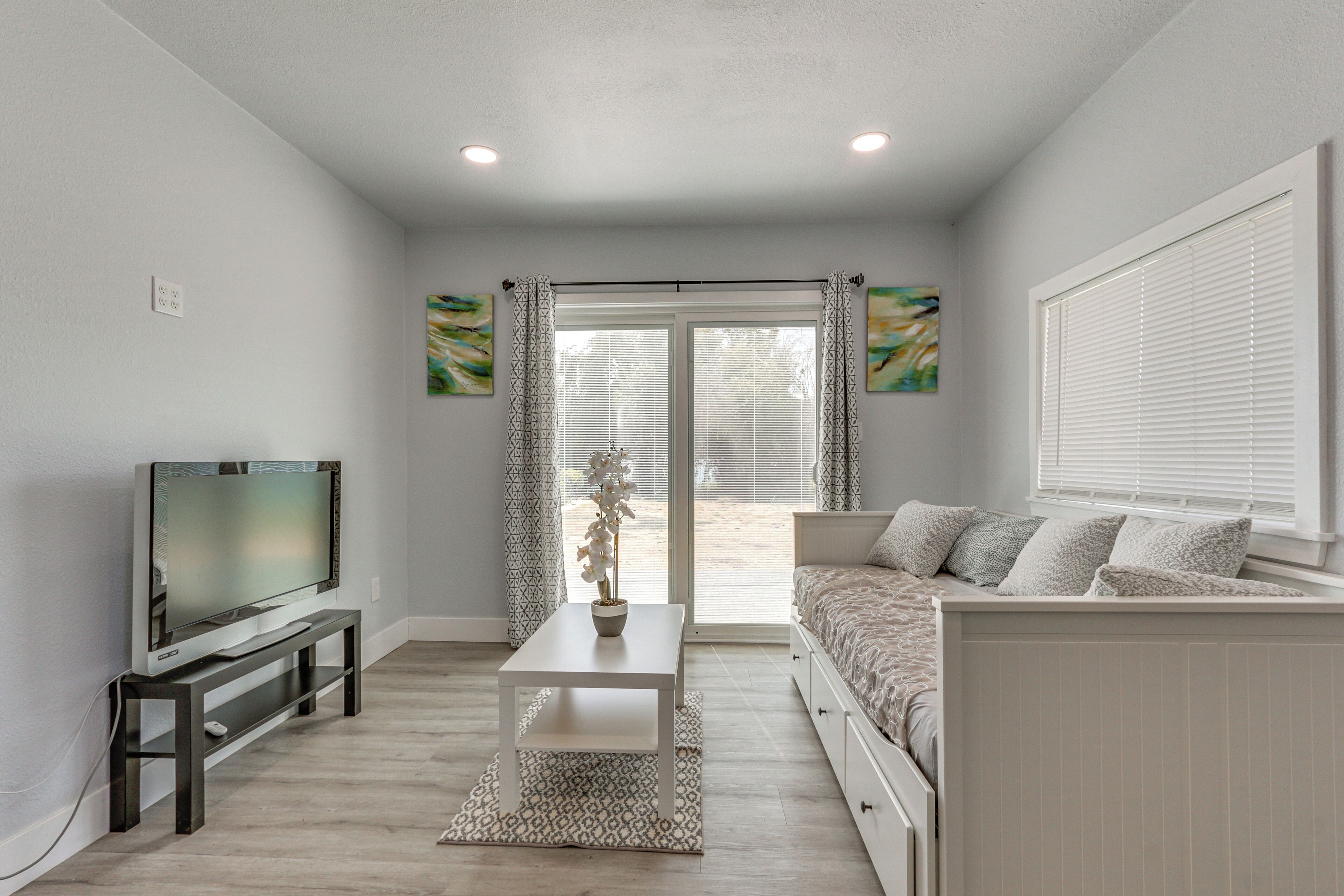 A modern living room with a flat-screen TV on a stand, a white coffee table, a sofa with patterned cushions, and a sliding glass door leading to an outdoor area.
