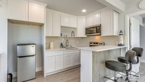 A modern kitchen with white cabinetry, a stainless steel refrigerator, a microwave, and a bar area with stools. - End Unit w/ Clear Lake Access in Norcal! (Clearlake)