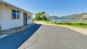 A single-story building with a black door and windows, a paved driveway, and a view of a lake and hills in the background. - End Unit w/ Clear Lake Access in Norcal! (Clearlake)