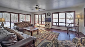 A spacious living room with a large sofa, a coffee table, and a television. There are large windows providing a view of the outdoors. - Snowmobile, Ice Fish & Explore! Lang Lake Cabin (Danbury)