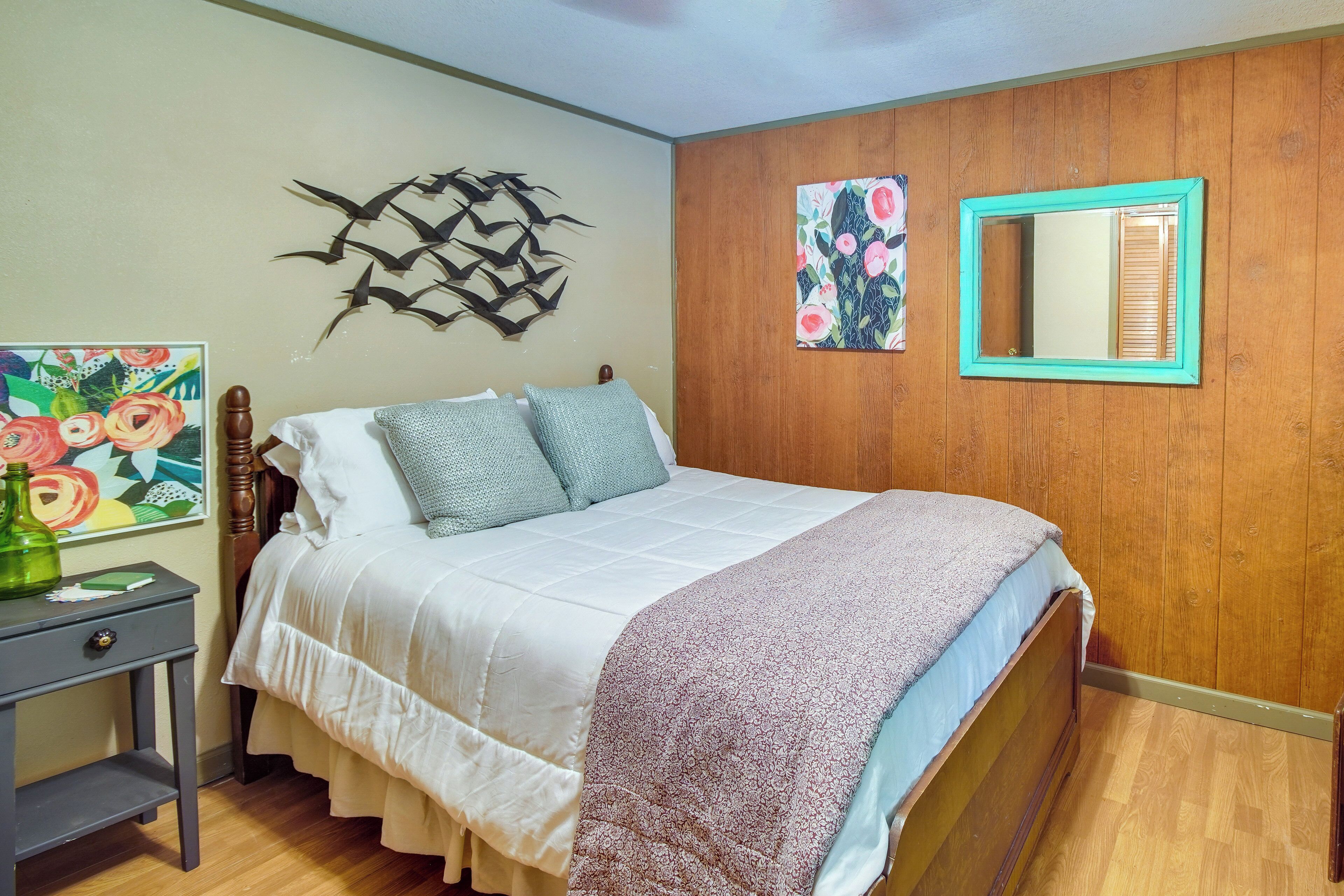 A bedroom with a wooden headboard, a bed with white bedding and a patterned throw, a nightstand with a green bottle, and wall art including a bird sculpture and a floral painting.