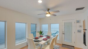 Un coin repas avec une table dressée pour quatre personnes, de grandes fenêtres, un ventilateur de plafond et une vue sur l’océan.