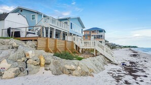 Une propriété située en bord de mer, comprenant une grande maison, une dépendance plus petite attenante et un escalier en bois menant à la plage.