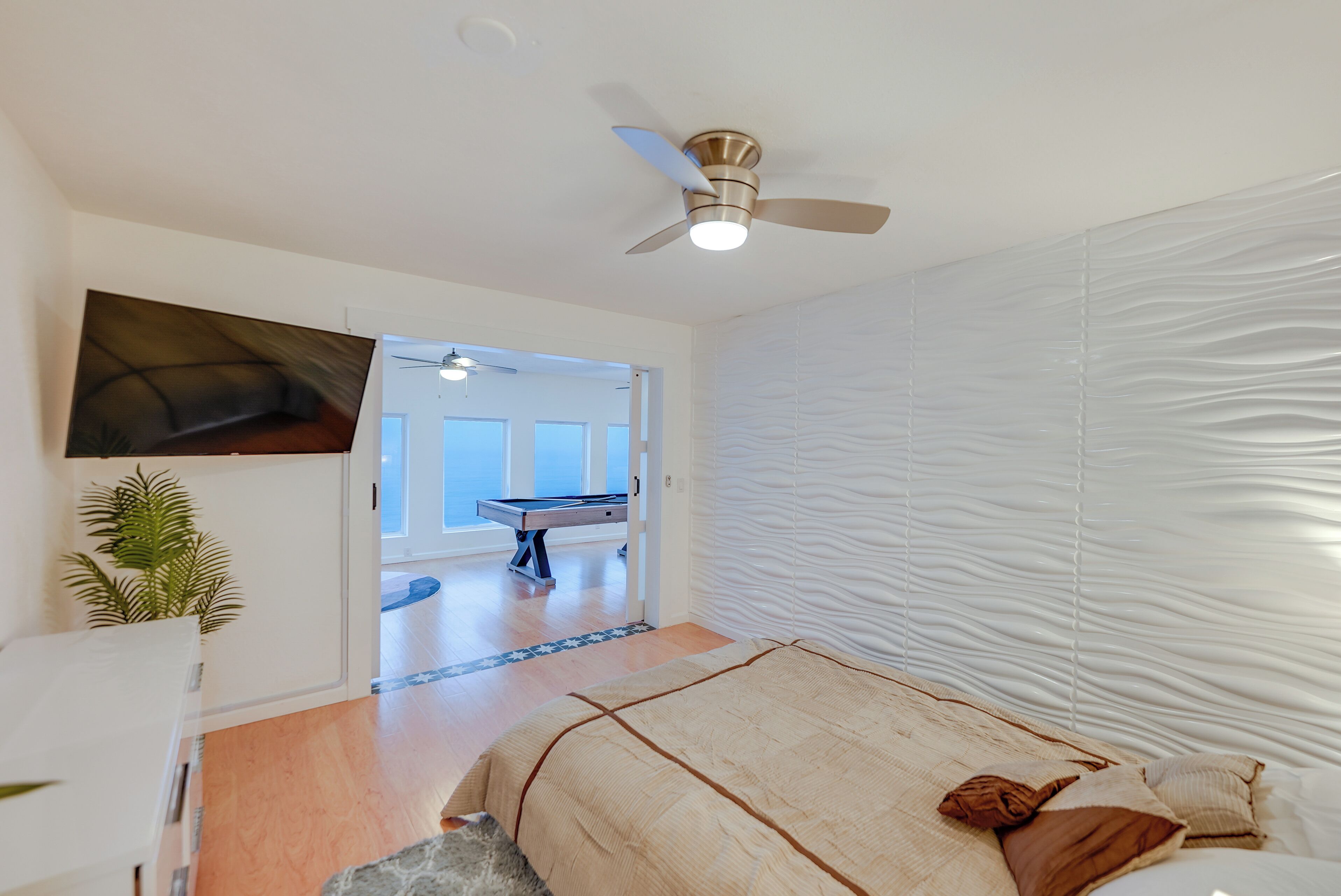 Une chambre avec un lit, un ventilateur de plafond, un téléviseur fixé au mur et une porte donnant sur un couloir où se trouve une table de billard.