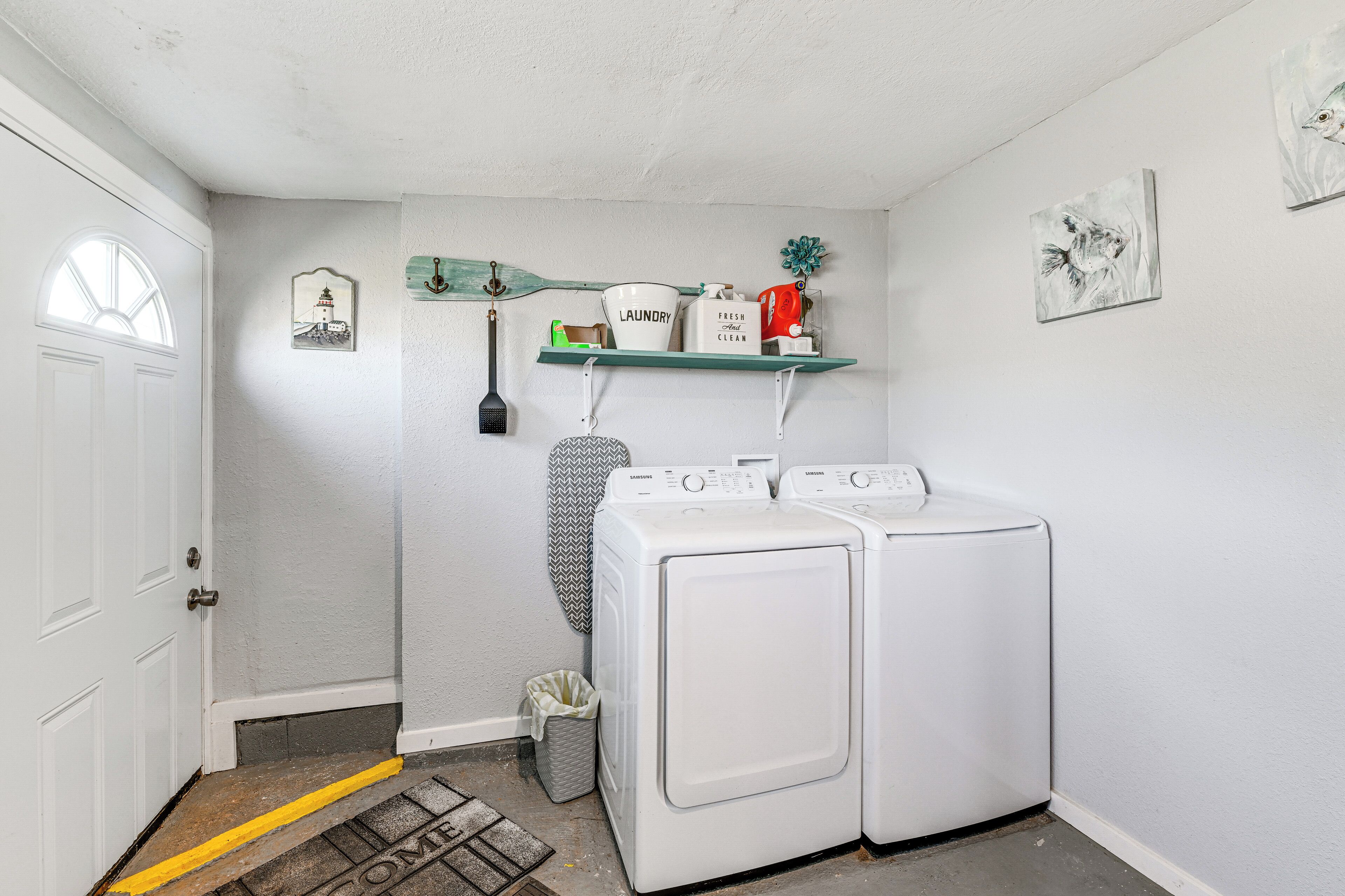 A laundry room with two white front-loading washers and dryers, a shelf with laundry supplies, a broom, and a trash can.