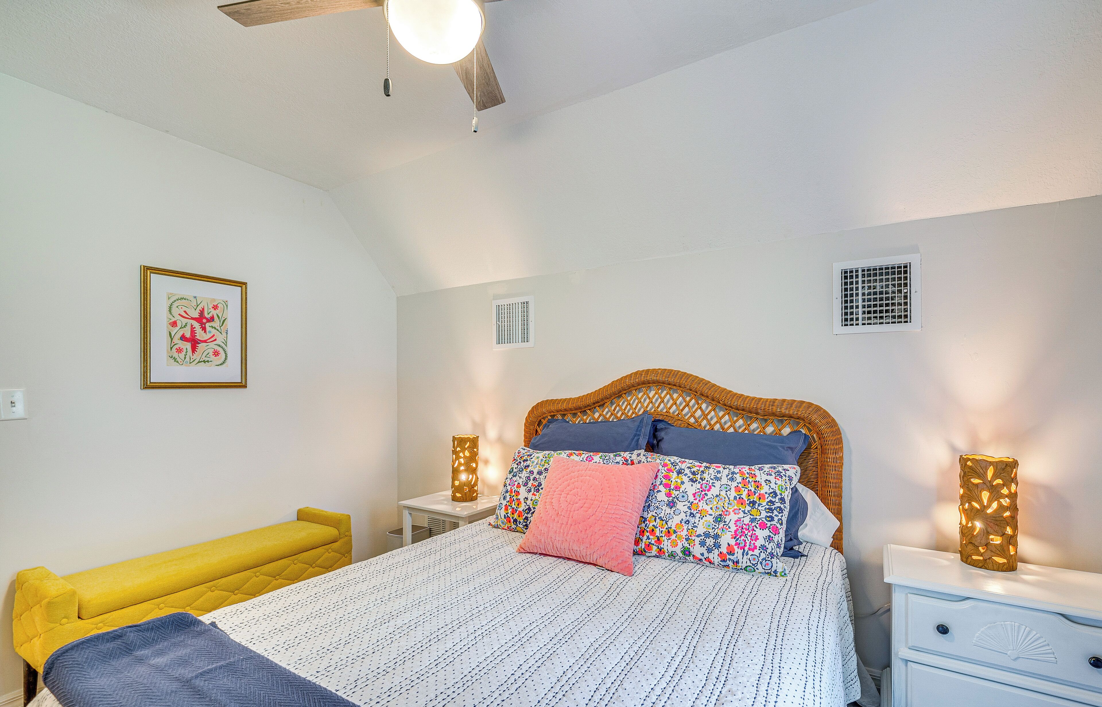 Un dormitorio con cama, mesitas de noche, un banco, un ventilador de techo, una lámpara de pared y una ventana.