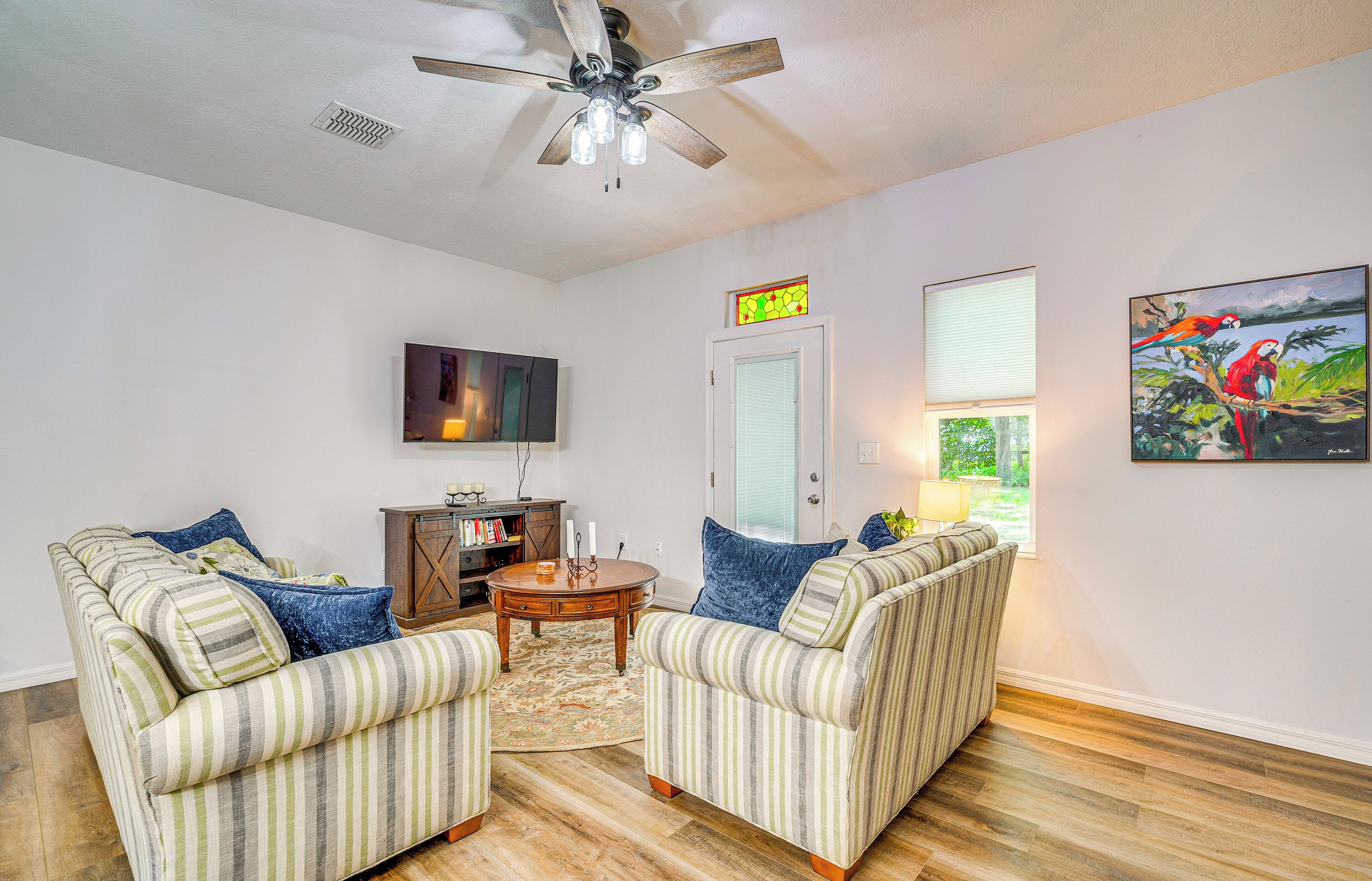A living room with striped sofas, a ceiling fan, a TV, a bookshelf, and a painting of parrots.