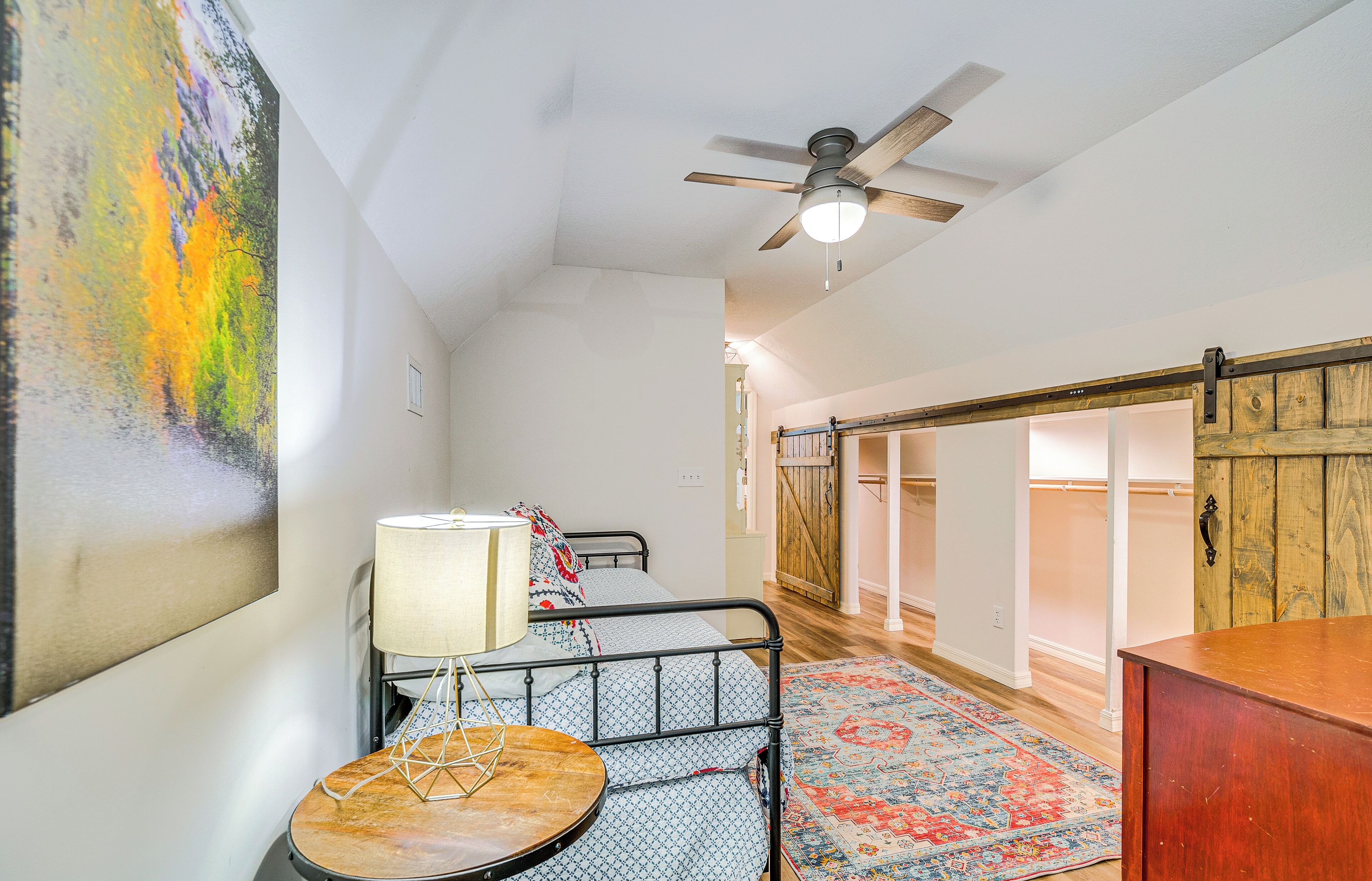 A bedroom with a bed, a nightstand, a lamp, a ceiling fan, and a large abstract painting on the wall.