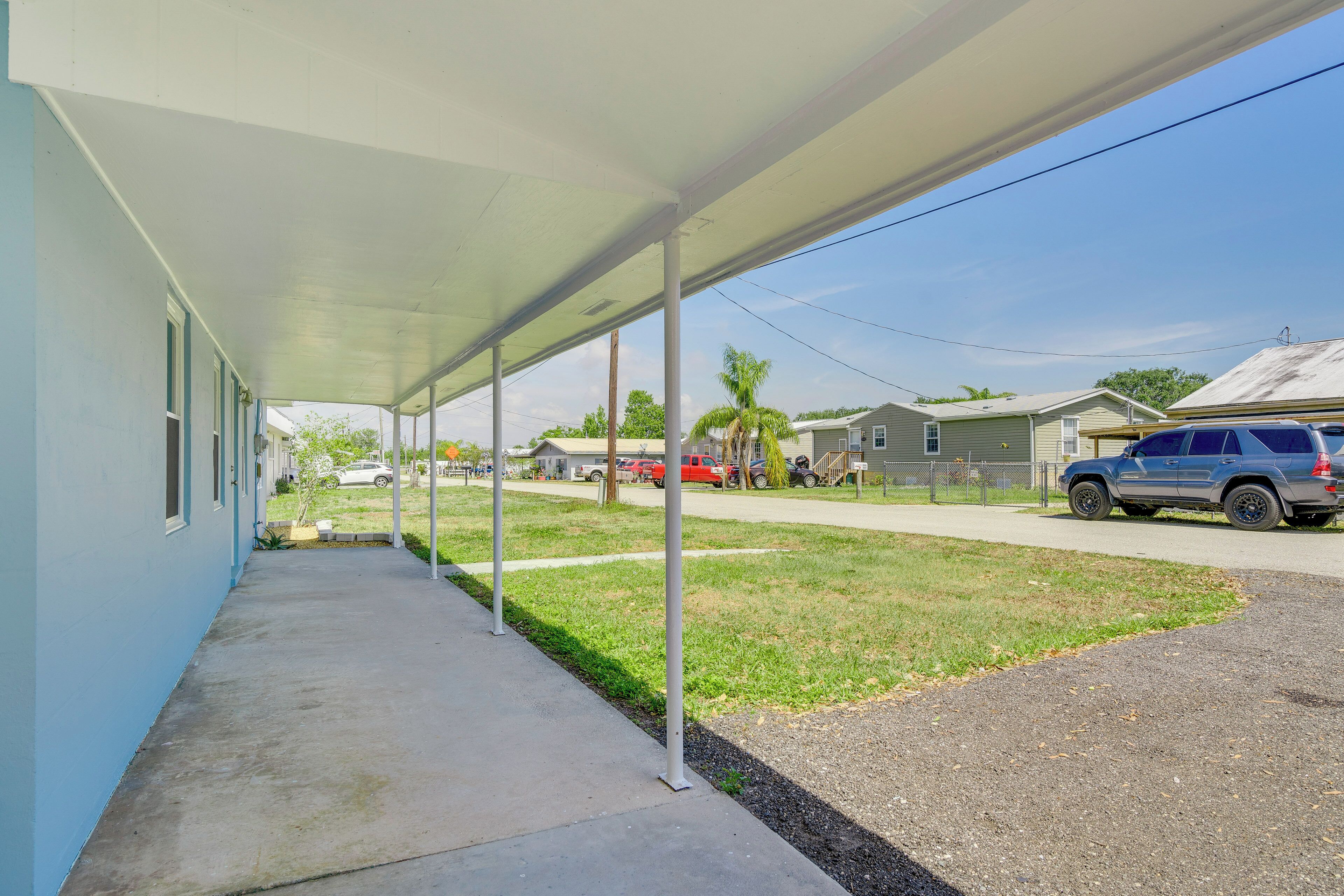 A covered outdoor area with a concrete walkway, metal columns, and a view of a street with parked cars and houses.
