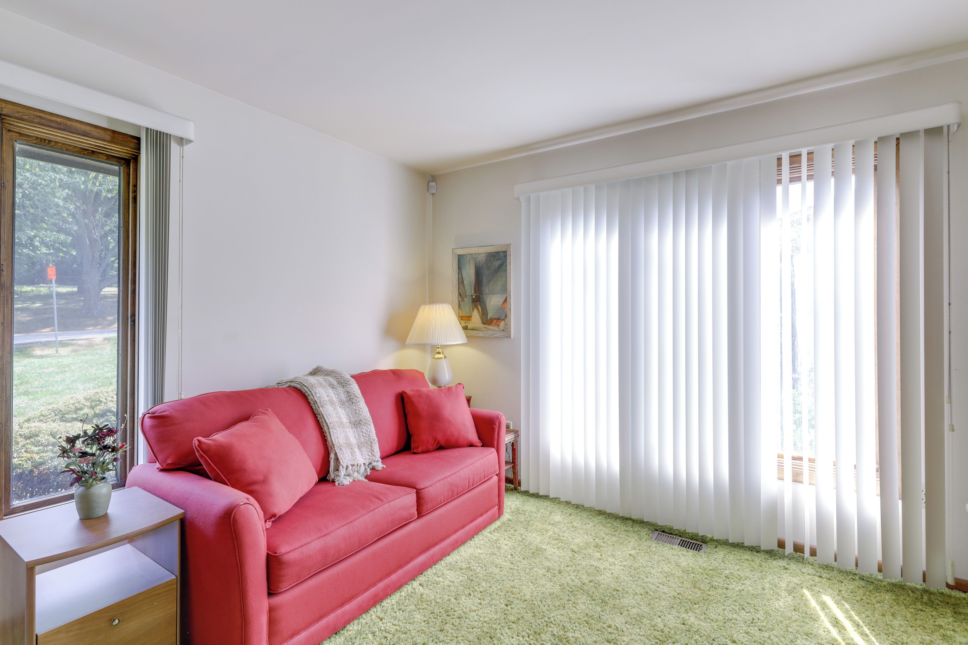 Une pièce avec un canapé rouge, une petite table, une lampe et une fenêtre avec des rideaux blancs.
