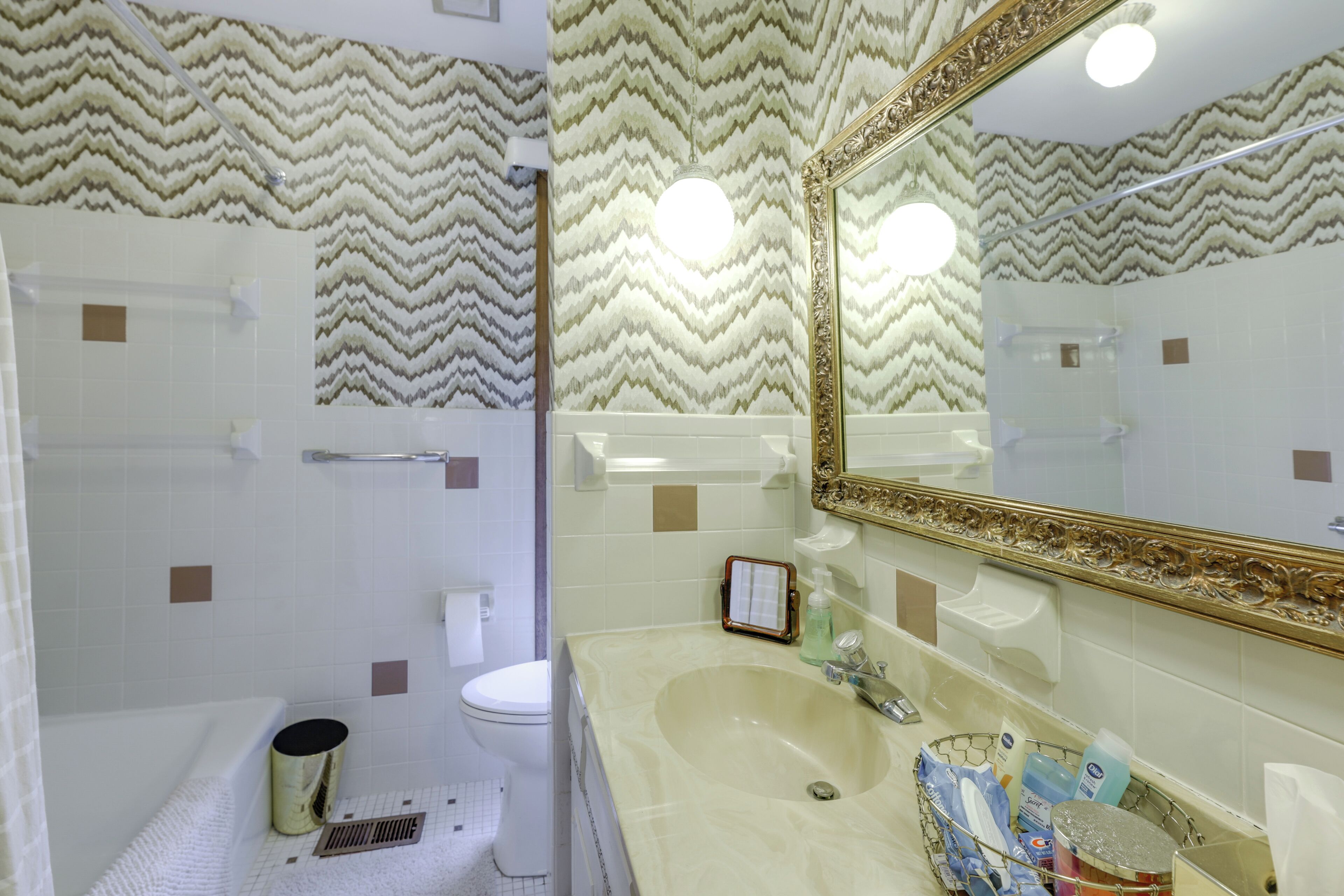 Baño con ducha, inodoro y lavamanos. Se observa un espejo con un marco ornamentado y artículos de tocador sobre el lavamanos.