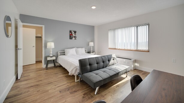 Quarto moderno com cama, mesas de cabeceira, sofá, janela com persianas e porta que dá para outro cômodo.