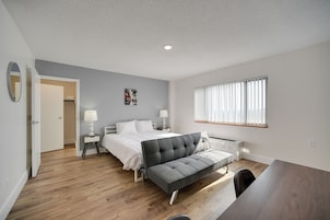 Un dormitorio moderno con cama, mesitas de noche, un sofá, una ventana con persianas y una puerta que da a otra habitación.