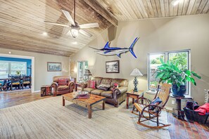 A spacious living room with a high ceiling, a ceiling fan, a large window with a view, a leather sofa, a wooden coffee table, and a rocking chair.