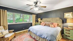Een slaapkamer met een groot bed, een plafondventilator, een raam met gordijnen, een nachtkastje en een stoel.
