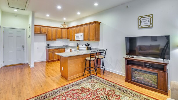 A modern kitchen with wooden cabinets, a central island with bar stools, a built-in microwave, and a flat-screen TV on a wooden stand. - 3 Mi to Warren Dunes: Sawyer Getaway w/ Fireplace! (Sawyer)