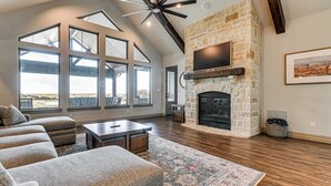 A spacious living room with a stone fireplace, large windows, and a ceiling fan. - Elegant 'sunset Meadow Ranch' in Texas w/ Hot Tub (Ponder)