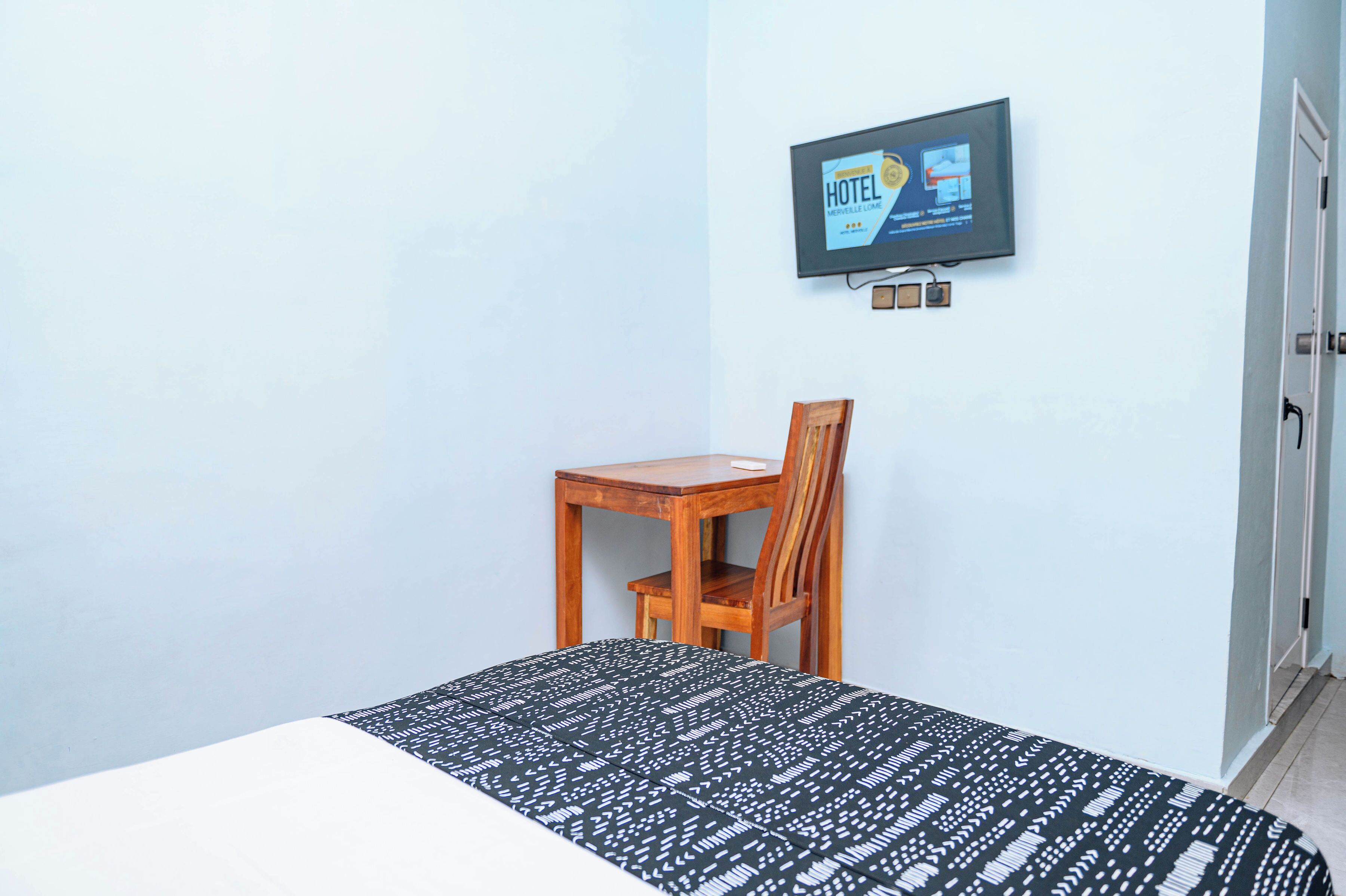 A hotel room with a bed, a wooden chair, a television mounted on the wall, and a door.