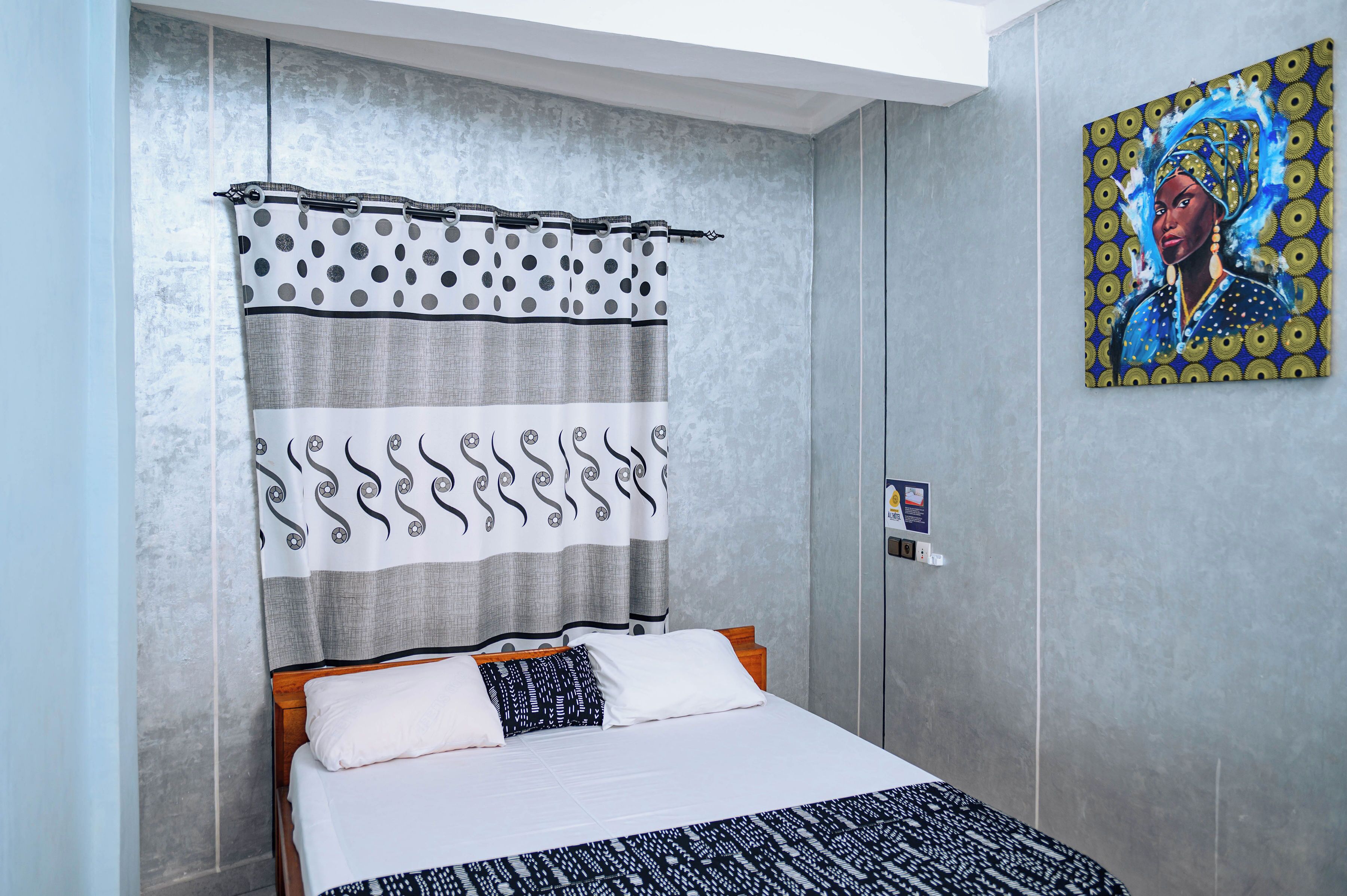 A hotel room with a bed, curtains, a painting on the wall, and a wall-mounted light switch.