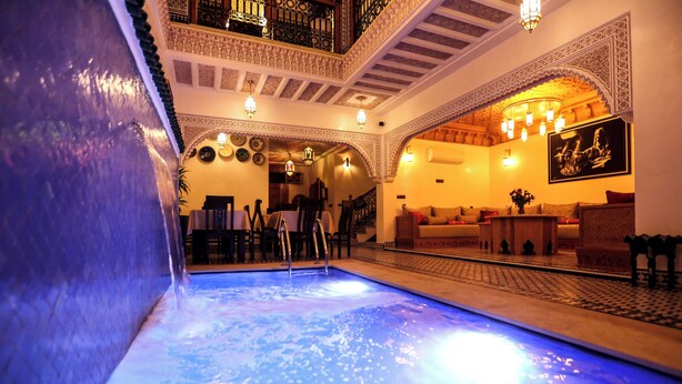 A luxurious indoor pool area with a blue-lit water feature, ornate architectural details, and a cozy seating area with cushions. - Riad Palais MERYEM (Marrakech)