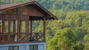 A wooden building with large windows and a balcony, surrounded by lush green hills. - Triptych by Xplore Indo (Kotagiri)