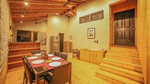 A wooden-floored dining area with a table set for four, a kitchen in the background, and a staircase leading to an upper level. - Triptych by Xplore Indo (Kotagiri)