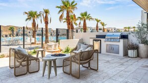 A rooftop patio with wicker furniture, a grill, and a view of a swimming pool and buildings.