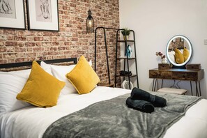 A neatly made bed with yellow and white pillows, a grey blanket, and a black towel. A round mirror with a lighted frame is on a wooden dresser beside the bed. - Charming London Apartment With Great Location (London)