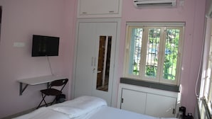 A bedroom with a bed, a desk, a chair, a TV, a wardrobe, and a window with a view of greenery. - Bonussa Homestay (Kolkata)