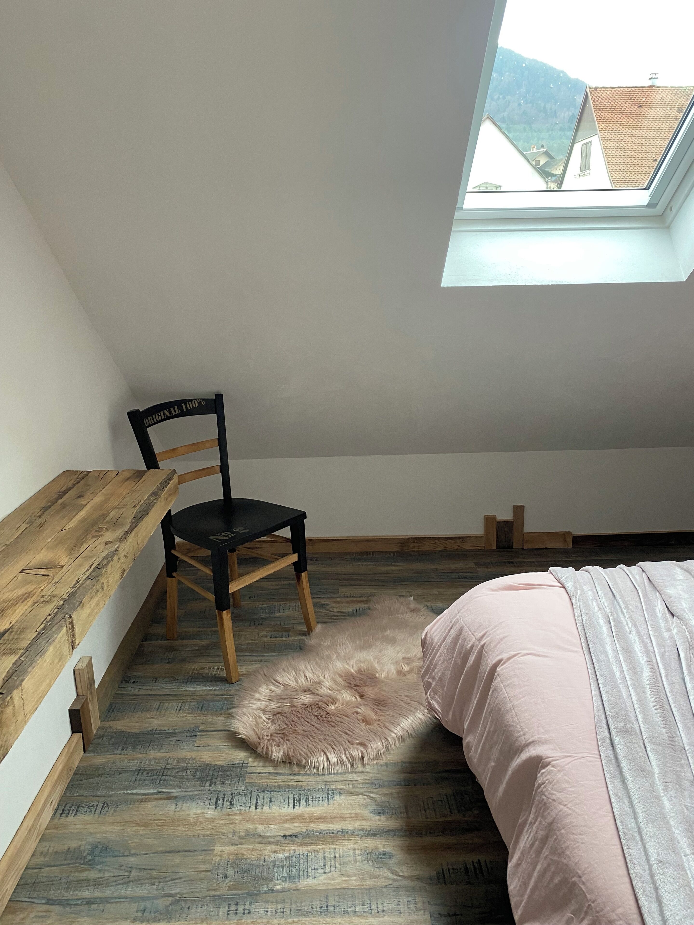 A room with a sloped ceiling, a skylight, a wooden desk, a chair, a fluffy rug, and a bed with a pink and white blanket.