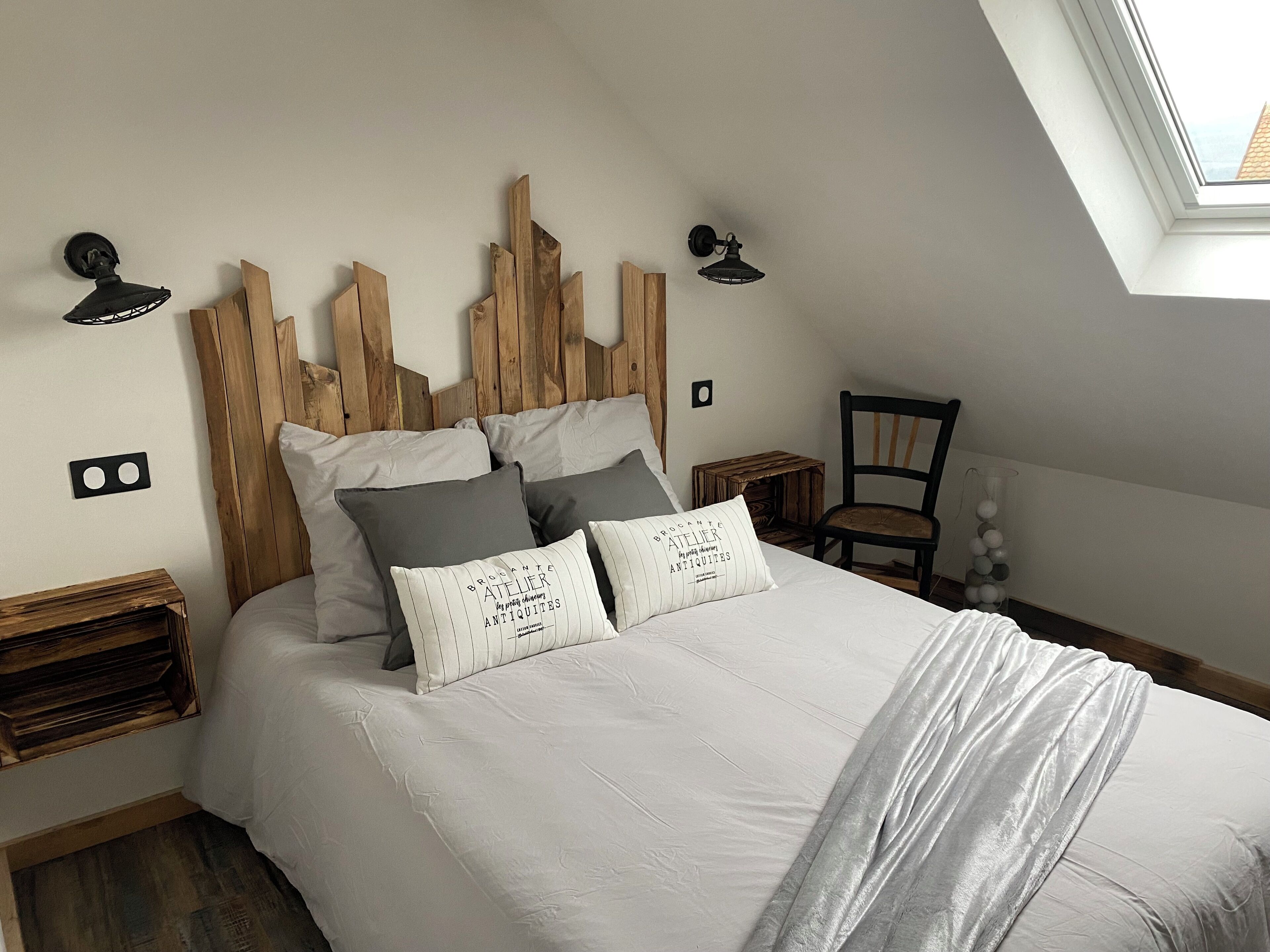 Ein Schlafzimmer mit schrägem Dach, einem Holzkopfteil, einem Bett mit weißer Bettwäsche, zwei Nachttischen, einem Sessel und einem Oberlicht.