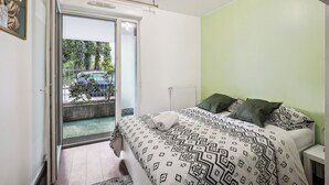 A bedroom with a bed, a radiator, a nightstand, and a view of a balcony with a car and greenery outside. - Apart-Chic-Les-Ulis with terrace and parking. (Les Ulis)