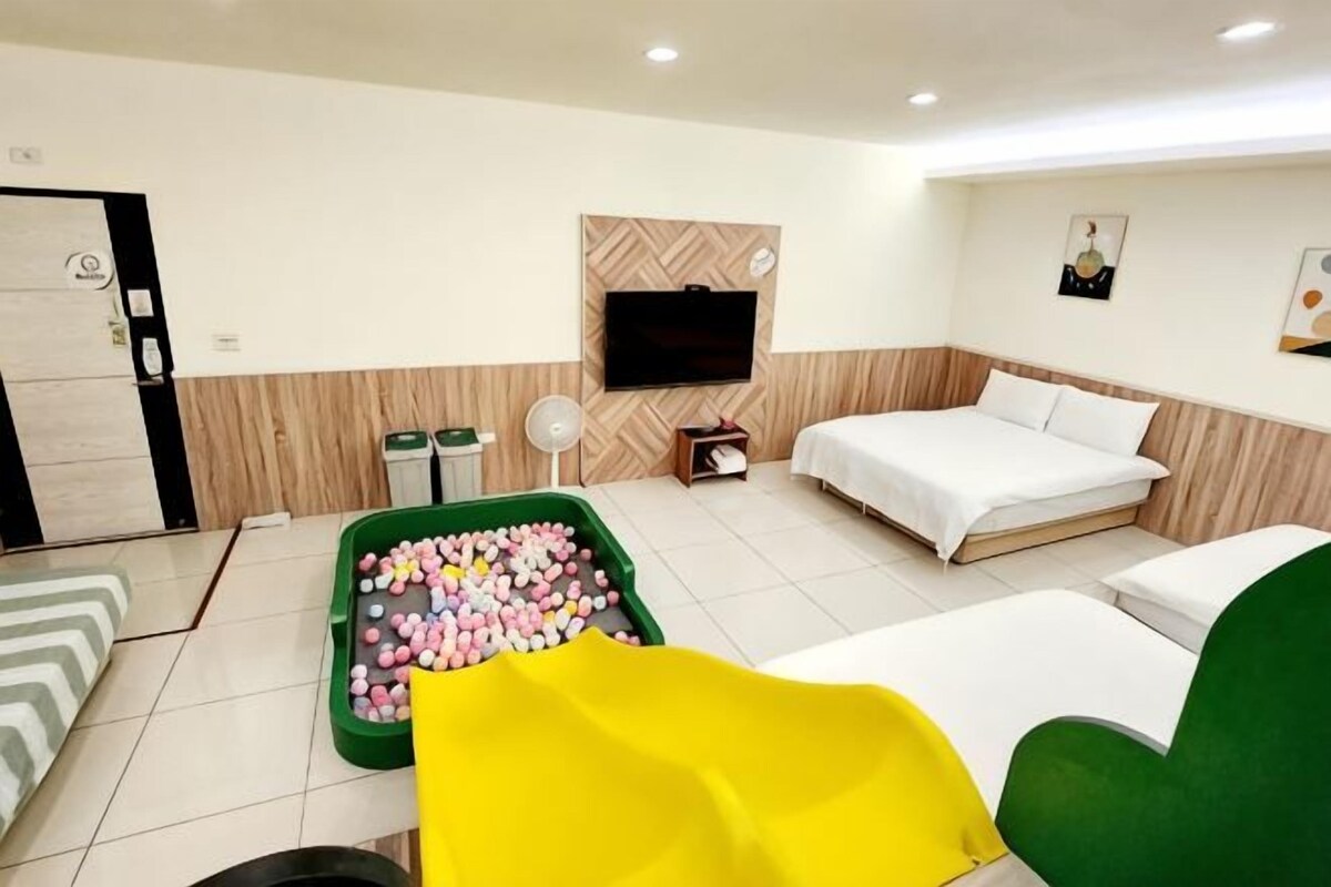 A hotel room with a bed, a green chair, a TV, a waste bin, and a trash bin.